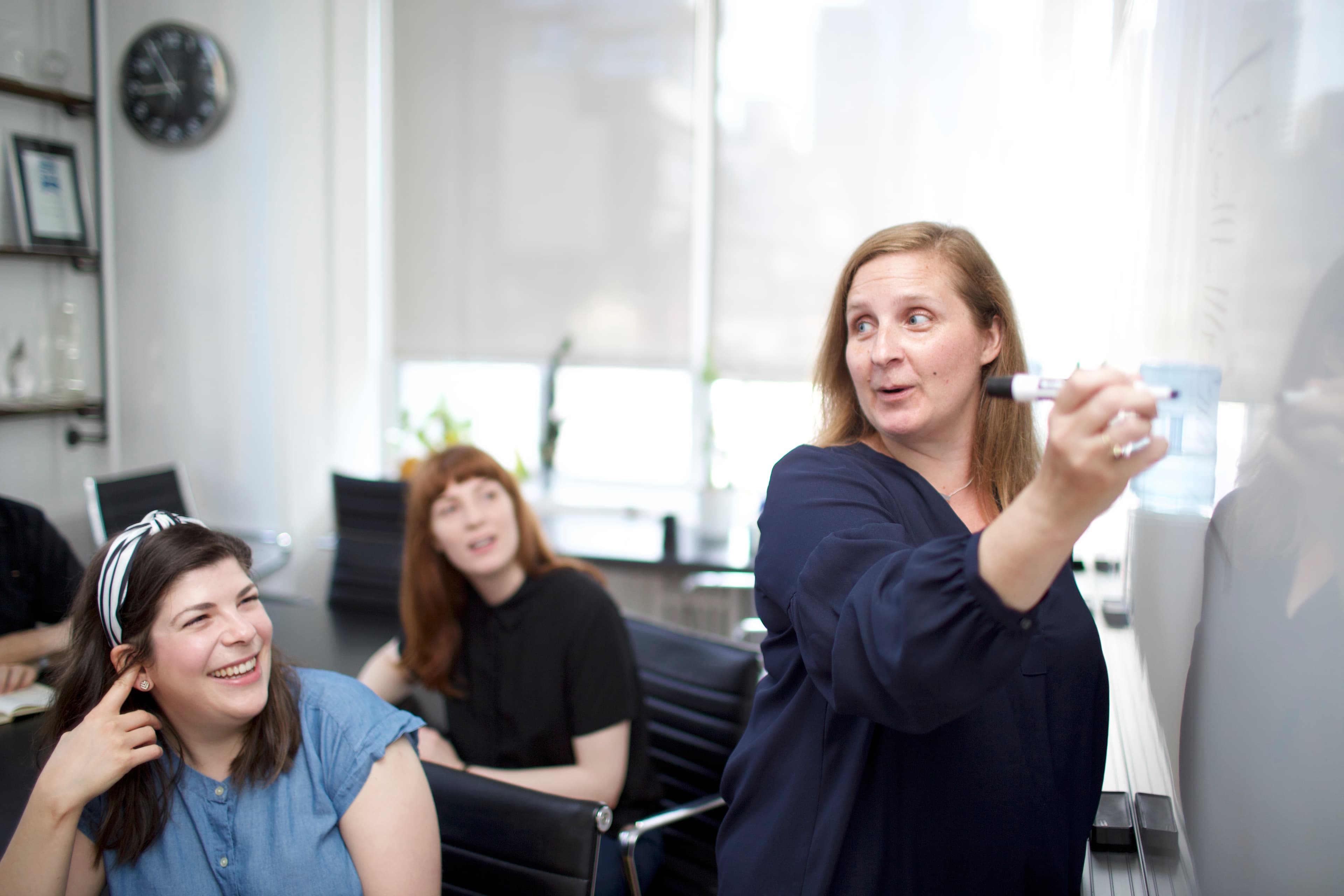 A woman stands at a white board with a marker in her hand. She's speaking and looking over her shoulder. Two women are sitting behind her, smiling and engaged.