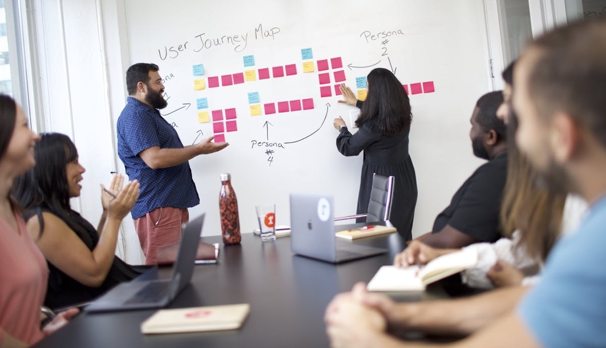 A diverse group of people are sitting at a conference table. At the front of the room, a man and woman gesture toward a whiteboard with colorful sticky notes on it.