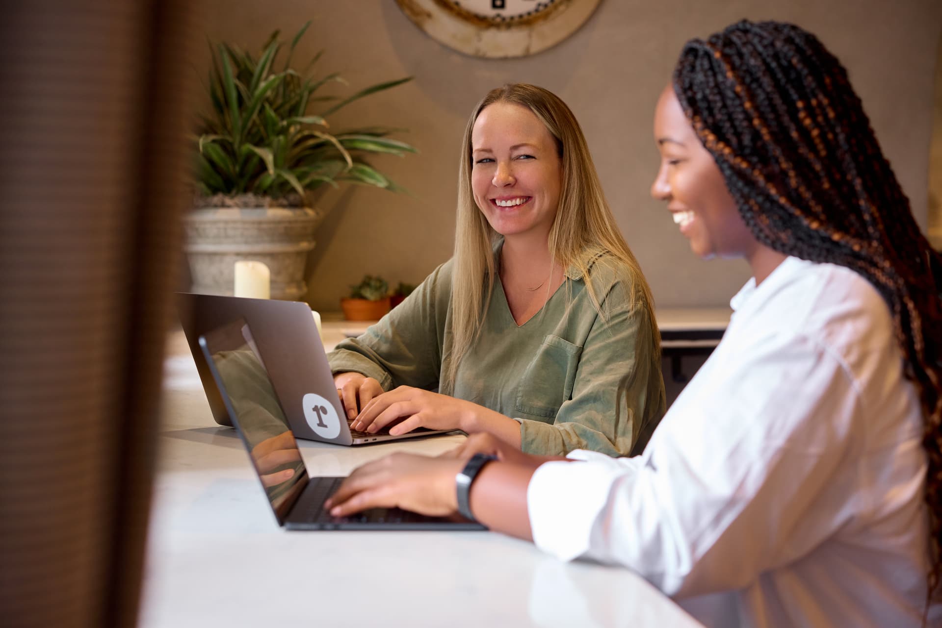Two women sit at a bar with laptops open and smiling