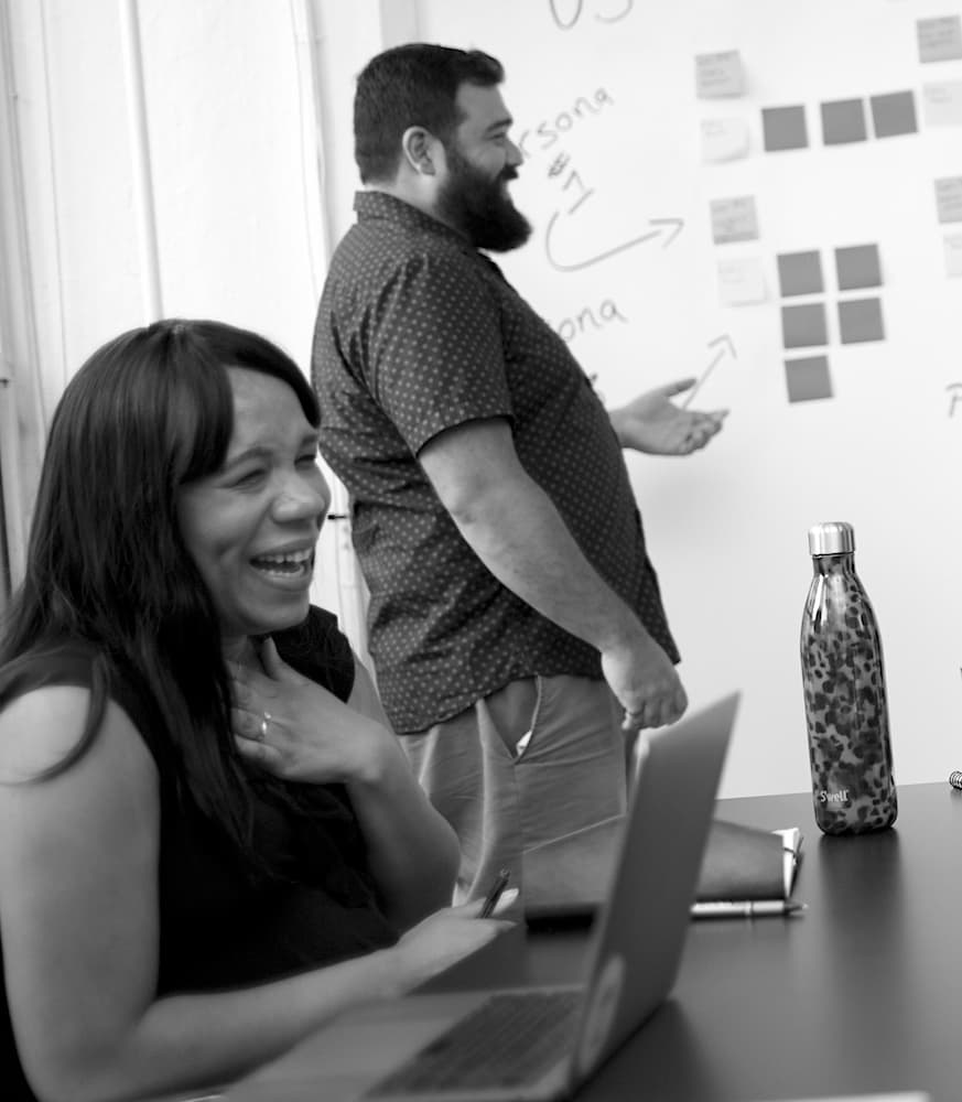 A woman is sitting in the foreground laughing. A man is standing in the background gesturing towards a whiteboard with sticky notes on it.