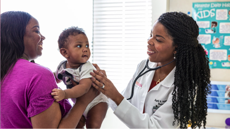 A Black mother is holding her baby while a Black female doctor examines her. Mother, doctor, and baby are all smiling.